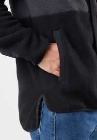 Giacca in fleece nera e grigia con superficie texturizzata, tasche laterali e orlo a contrasto. Il design presenta una vestibilità comoda e linee eleganti.