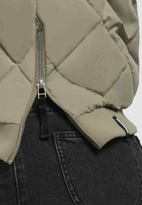 Khaki gewatteerde jas met een rits en een zwarte elastische boord, gecombineerd met donkere jeans. Beschikt over een textuurstof en stiksels benadrukken.