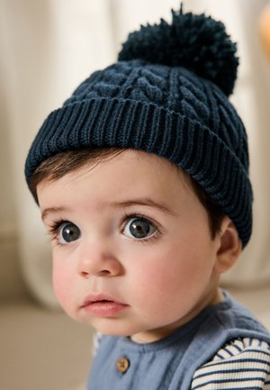 Bambino con grandi occhi che indossa un cappello lavorato a maglia scuro con pon pon e un completo blu con bottoni e maniche a righe, che guarda leggermente di lato.