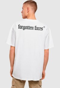 Balta per didelį t-shirt, pagaminta iš medvilnės, su juodu gotikiniu tekstu "pamesti veidai" ant viršutinės nugaros dalies. Trumpomis rankovėmis, laisvas kirpimas.