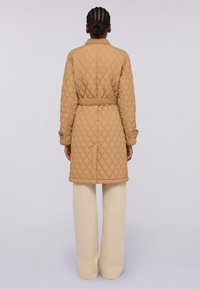Cappotto trapuntato beige con cintura in vita, maniche lunghe e colletto, indossato sopra pantaloni a palazzo crema. Il cappotto presenta un motivo a diamante.