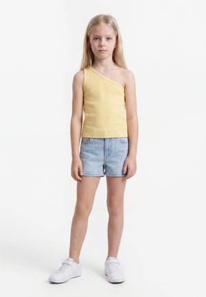 Jong meisje staand, draagt een geel topje met één schouder, lichtblauwe spijkershorts en witte sneakers tegen een effen witte achtergrond.