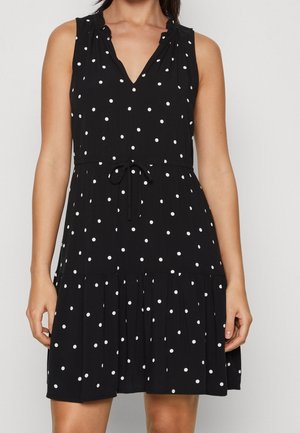 Czarna sukienka bez rękawów w białe kropki, z dekoltem w serek, talią ze sznurkiem i falbanką na dole, noszona przez stojącą osobę.