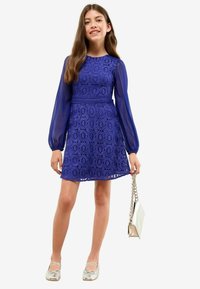Ragazza che indossa un abito di pizzo blu con maniche lunghe trasparenti, ballerine argentate e tiene una borsa argentata con una tracolla a catena.