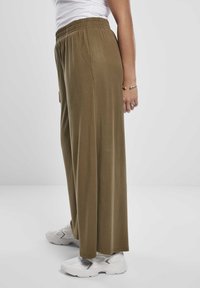 Personne portant un pantalon ample vert olive, des baskets blanches et un haut blanc, debout de profil sur un fond gris neutre.