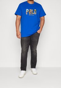 Blå bomullströja med en grafisk "POLO"-tryck i färgglada mönster; i kombination med mörka jeans och vita sneakers, stående på en neutral bakgrund.