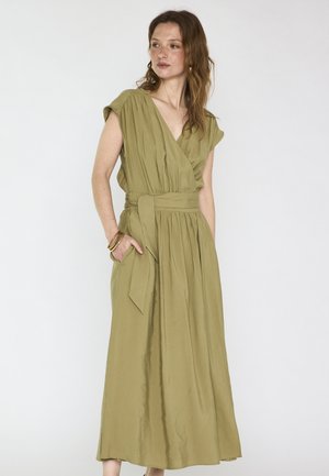 Vrouw met golvend bruin haar, gekleed in een mouwloze olijfgroene midi-jurk met een V-hals en tailleband, poserend met één hand in de zak tegen een witte achtergrond.