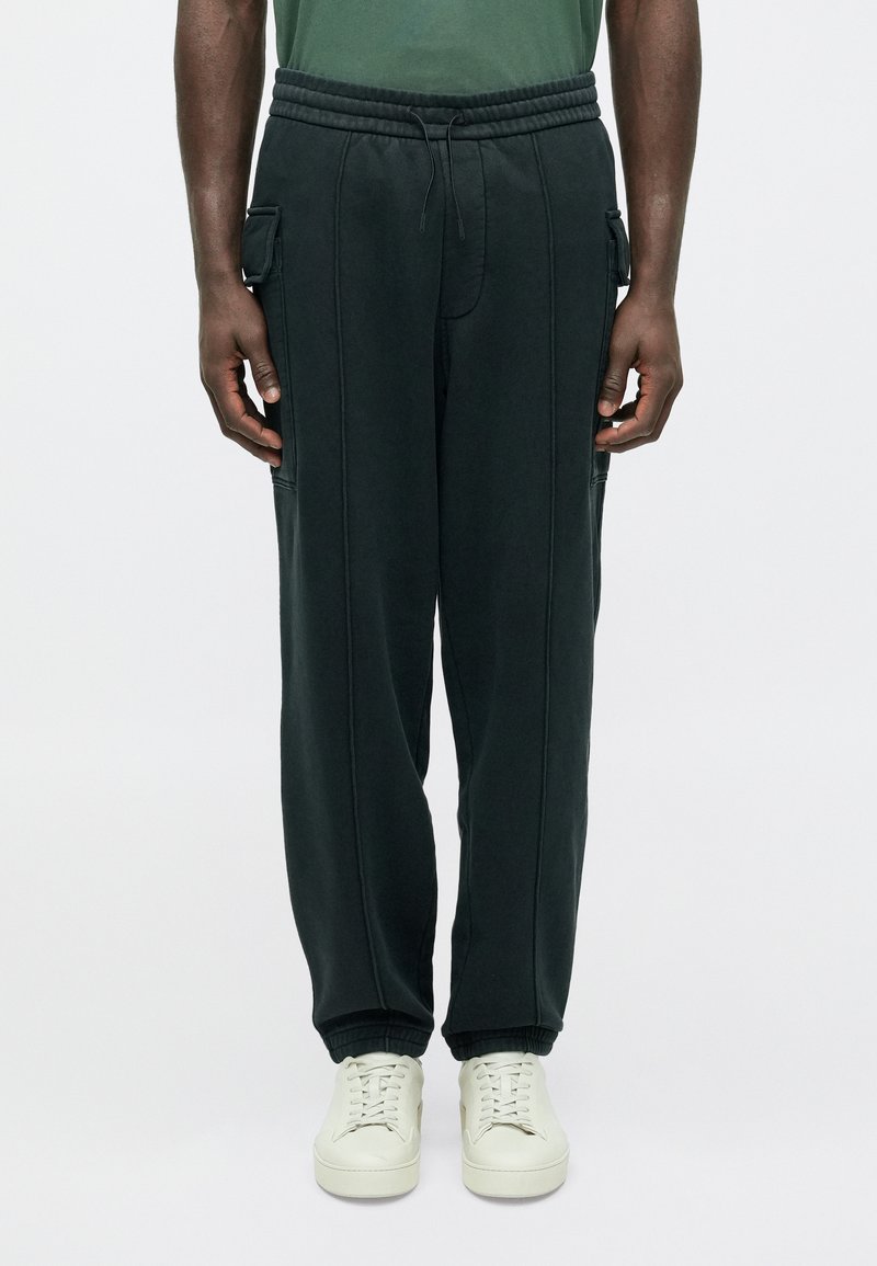 Tmavozelené tepláky s elastickým pásom, bočnými vreckami a zúženými manžetami. Vyrobené z mäkkej látky s viditeľnými švami ako dekoráciou.