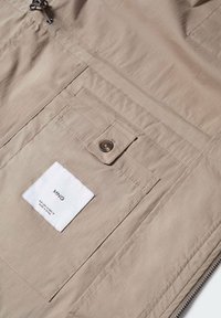 Veste beige à la texture lisse, dotée d'une poche avant, d'une fermeture à boutons et d'une étiquette tissée avec le logo de la marque. Détail de conception avec fermeture éclair inclus.