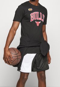 Sort T-shirt med teksten "BULLS" og logo, parret med sort-hvide mesh-basketballshorts. Holder en brun basketball og har en sort sweatshirt på.