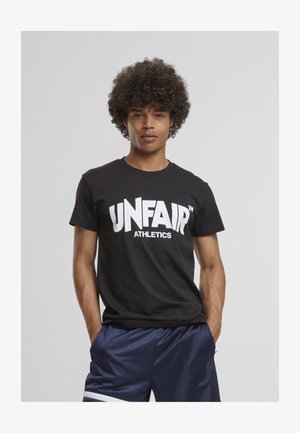 Junger Mann mit lockigem Haar, der ein schwarzes "UNFAIR ATHLETICS" T-Shirt und marineblaue Sportshorts trägt, steht mit den Händen in den Taschen.