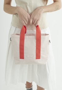 Lyserød tote bag med røde båndhåndtag, rektangulær form, struktureret stof, lille logo på forsiden, og metalaccenter på siderne.