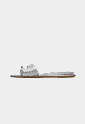 Επίπεδη ασημί μεταλλική σαγιονάρα με μία φαρδιά λωρίδα και θηλιά για τα δάχτυλα, σε ανοιχτό καφέ σόλα, απεικονιζόμενη από το πλάι σε λευκό φόντο.