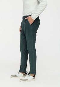 Sötétzöld kordbársony nadrágot viselő férfi, aki a zsebeiben tartja a kezét, fehér sportcipőben áll egy világos, egyszínű háttér előtt.