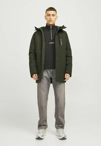 Grøn polstret jakke med hætte, lynlås lukning og logo detalje, båret over en sort bluse og parret med grå jeans og sølv sneakers.