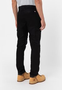 Man wearing black cargo pants and tan lace-up work boots standing on white background, viewed from behind.