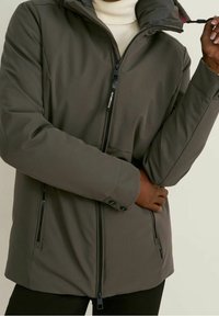 Šedá zateplená bunda s vysokým límcem, předním zipovým uzávěrem a dvěma bočními kapsami na zip. Má manžety na knoflíky a měkkou texturu.