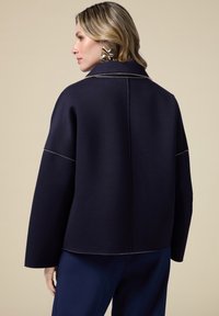 Giacca blu navy con un design strutturato, caratterizzata da un colletto alto, maniche larghe e un leggero piping lungo le cuciture. Tessuto morbido.