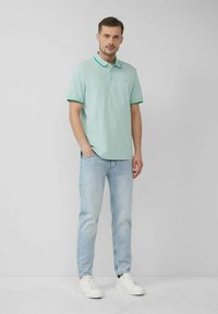 Mintgrünes Poloshirt mit kontrastierendem türkisfarbenem Kragen, kombiniert mit hellblauen Jeans und weißen Sneakers. Einfaches Design, kurze Ärmel.