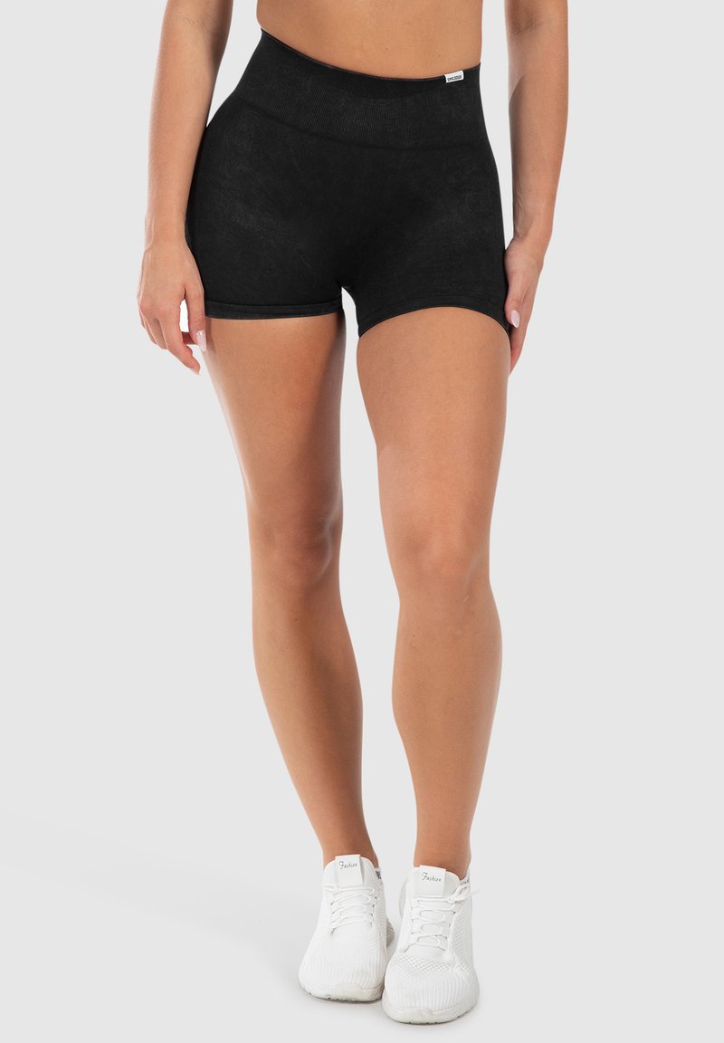 Zwarte sportshorts met een hoge tailleband, een gladde textuur en een korte snit. Draagt witte sneakers. Effen achtergrond.