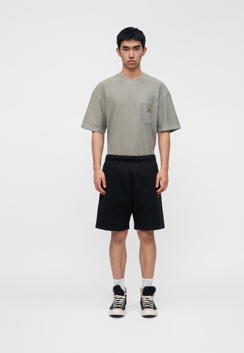 Junger Mann steht, trägt ein graues oversized T-Shirt, schwarze Shorts, weiße Socken und schwarze High-Top-Sneaker vor hellem einfarbigem Hintergrund.