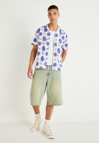 Majica kratkih rukava u bijeloj boji s plavim grafičkim uzorkom, uparena s kratkim trapericama svijetle boje i visokim tenisicama. Casual fit.