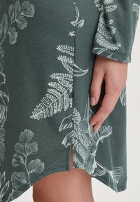 Abito verde con stampe botaniche bianche, caratterizzato da uno spacco laterale e maniche lunghe. Realizzato in un tessuto morbido e testurizzato.