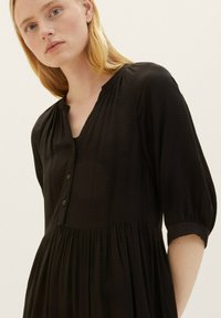Zwarte blouse jurk met een v-hals, textuurstof, driekwart mouwen en een uitlopende rok. Bevat rimpeling op de taille.