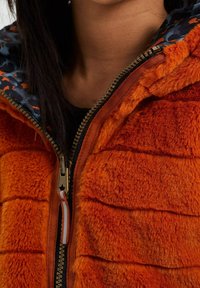 Orange faux pälsjacka med kviltad textur, som har ett mönstrat marinblått foder och en guldfärgad dragkedja med en stjärndetalj på dragfliken.