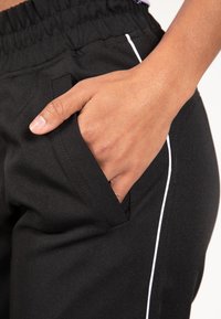 Zwarte sportbroek met een elastische tailleband, voorzien van een zijzak en witte bies langs de buitennaad. Een gladde stoftextuur.