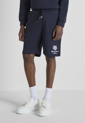 Modello maschile che indossa pantaloncini con coulisse blu navy con logo bianco sulla coscia, calzini bianchi alla caviglia e scarpe da ginnastica bianche, in piedi contro uno sfondo grigio chiaro.