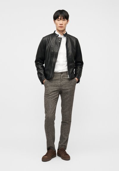 Άνδρας που στέκεται με τα χέρια στις τσέπες, φοράει μαύρο δερμάτινο μπουφάν, λε υκό πουκάμισο, γκρι παντελόνια και καφέ αθλητικά παπούτσια, μπροστά σε απλό φόντο.