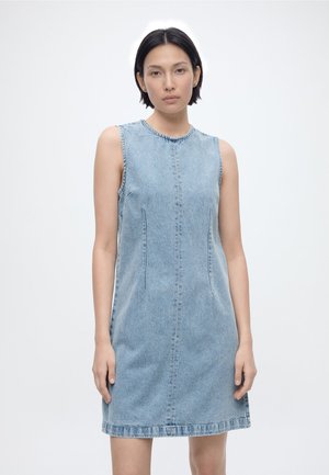 Γυναίκα με κοντά σκούρα μαλλιά που φοράει αμάνικο ανοιχτό γαλάζιο φόρεμα από ντένιμ, στέκεται μπροστά σε λευκό φόντο.