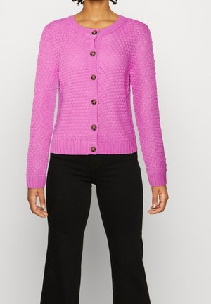 Cardigan rosa lavorato a maglia con un motivo testurizzato, scollatura rotonda e sette bottoni marroni. Maniche e orlo aderenti, abbinato a pantaloni neri a zampa.