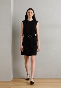 Vestido negro sin mangas con un diseño ajustado, cinturilla ceñida y hebilla de cinturón circular. La modelo lo combina con zapatos planos negros sobre un suelo de madera.