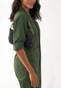 Sac à main en cuir noir avec une bandoulière en chaîne dorée, présentant un design incurvé élégant et des accents en bouton. Porté par-dessus une combinaison vert olive.