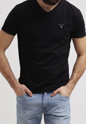 Man wearing black V-neck GANT t-shirt and light blue jeans with hands in pockets against plain light background.