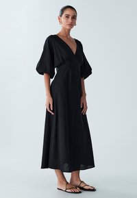 Must kleit, mille pikkus ulatub põlvedeni, sügava V-kaelusega, paksude varrukatega ja voolava seelikuga. Pehme kangas, sileda tekstuuriga, kantud minimalistlike sandaalidega.