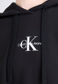 Felpa nera con cappuccio realizzata in materiale morbido, caratterizzata da un logo bianco di Calvin Klein Jeans. Include lacci regolabili e un design classico.