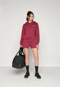 Hanorac maroniu cu șnur și logo, pantaloni scurți maronii asortati, geantă neagră, încălțări negre și șosete negre, stând pe un fundal simplu.