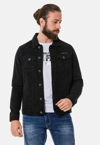 Veste en denim noir avec un design col, deux poches poitrine, des boutons en métal, des manches longues et une coupe légèrement cintrée. Portée par-dessus une chemise blanche.