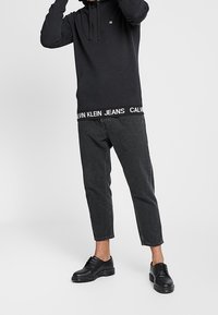 Osoba v černé mikině Calvin Klein a zkrácených černých džínech s černými koženými botami, stojící proti jednobarevnému světlému pozadí.