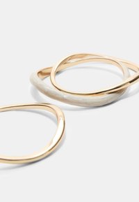Anelli intrecciati oro e crema; uno è in metallo dorato lucido, l'altro ha una finitura lucida con una texture e una sottile marmorizzazione.