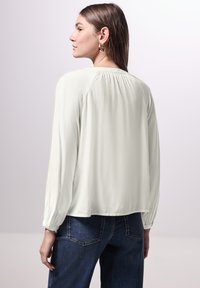 Lichtbeige blouse met lange mouwen en geplooide details aan de achterkant, gecombineerd met donkerblauwe spijkerbroek, gladde textuur, relaxte pasvorm.