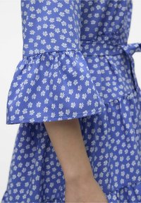 Niebieska sukienka w kwiaty z falbaniastym rękawem, wykonana z bawełnianego materiału w małe białe wzory kwiatowe, z detalem w postaci wiązania w talii.