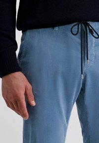 Man in een lichtblauwe corduroybroek met zwarte trekkoorden en een zwarte lange mouwtrui, met een ontspannen hand langs de zijde.