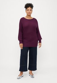 Lila gestrickter Pullover mit weitem Halsausschnitt und Dreiviertelärmeln, kombiniert mit weit geschnittenen, dunklen Jeans und silbernen flachen Schuhen.