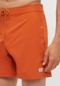 Narancssárga fürdőshorts sima anyagból, húzózsinórral ellátott derékkal, oldalsó zsebekkel és egy kis márkás címkével a szegély közelében.