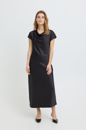 Fiatal nő fekete, selymes, rövid ujjú, bokáig érő ruhában és fekete hegyes orrú magassarkú cipőben, egy egyszerű, világos háttér előtt áll.
