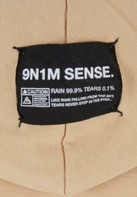 Etichetta in tessuto beige con testo nero che riporta "9N1M SENSE," dettagliando la resistenza alla pioggia. Include simbolo di cautela e istruzioni per la cura, con texture visibile.